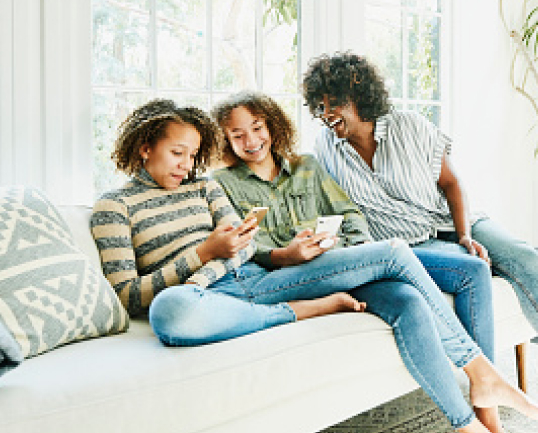Three women ranging in age sit on a couch using cellphones.